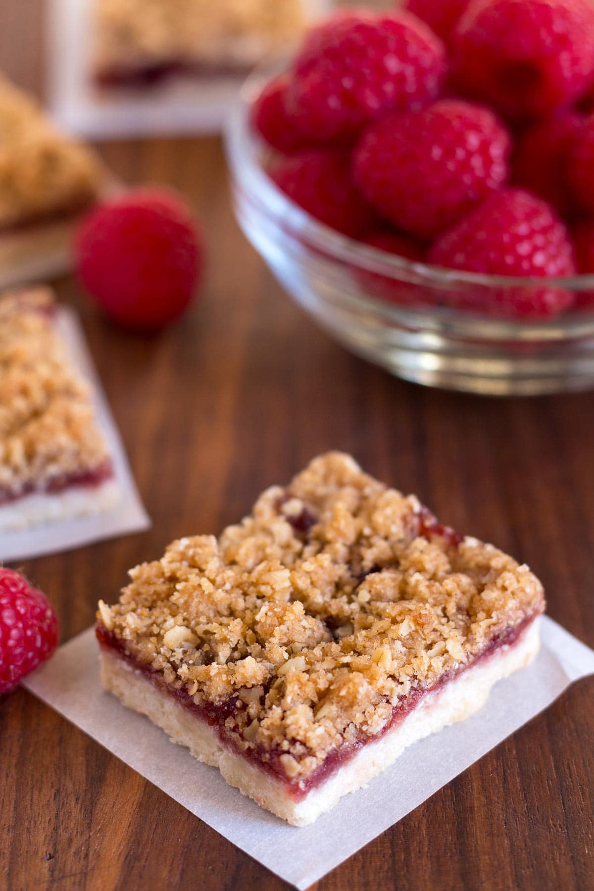 Raspberry bars with oat topping on parchment square with fresh raspberries in the background.