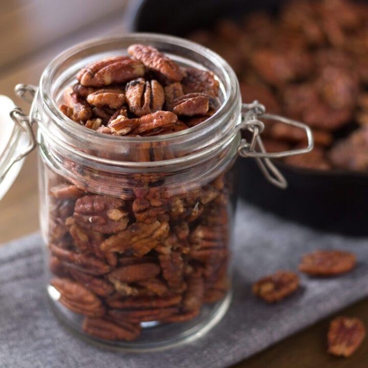 Open jar of maple pecans with cast iron pan in background