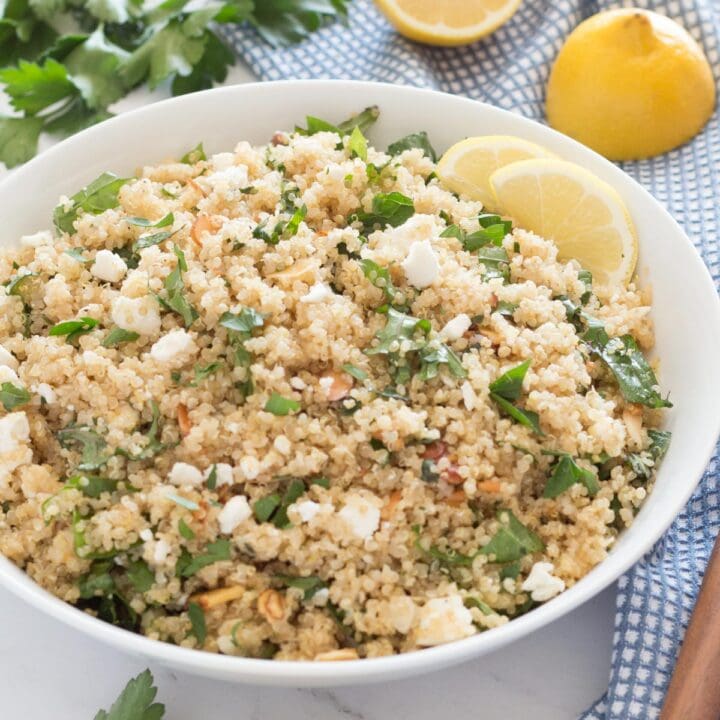 Bowl of lemon quinoa salad with feta and herbs next to blue towel and wooden spoon.