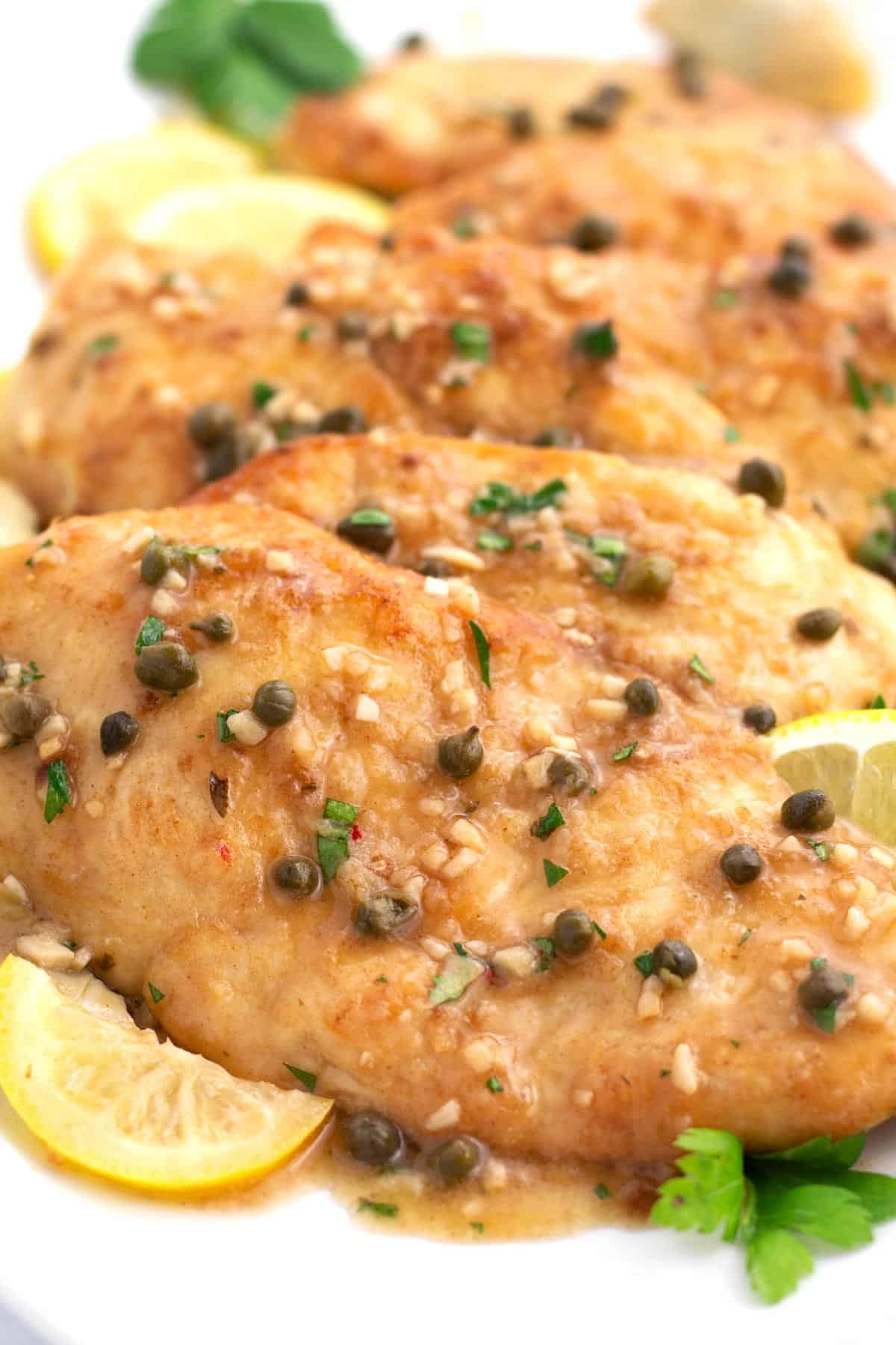 White platter with pieces of chicken piccata sprinkled with capers, parsley, and fresh lemon slices.