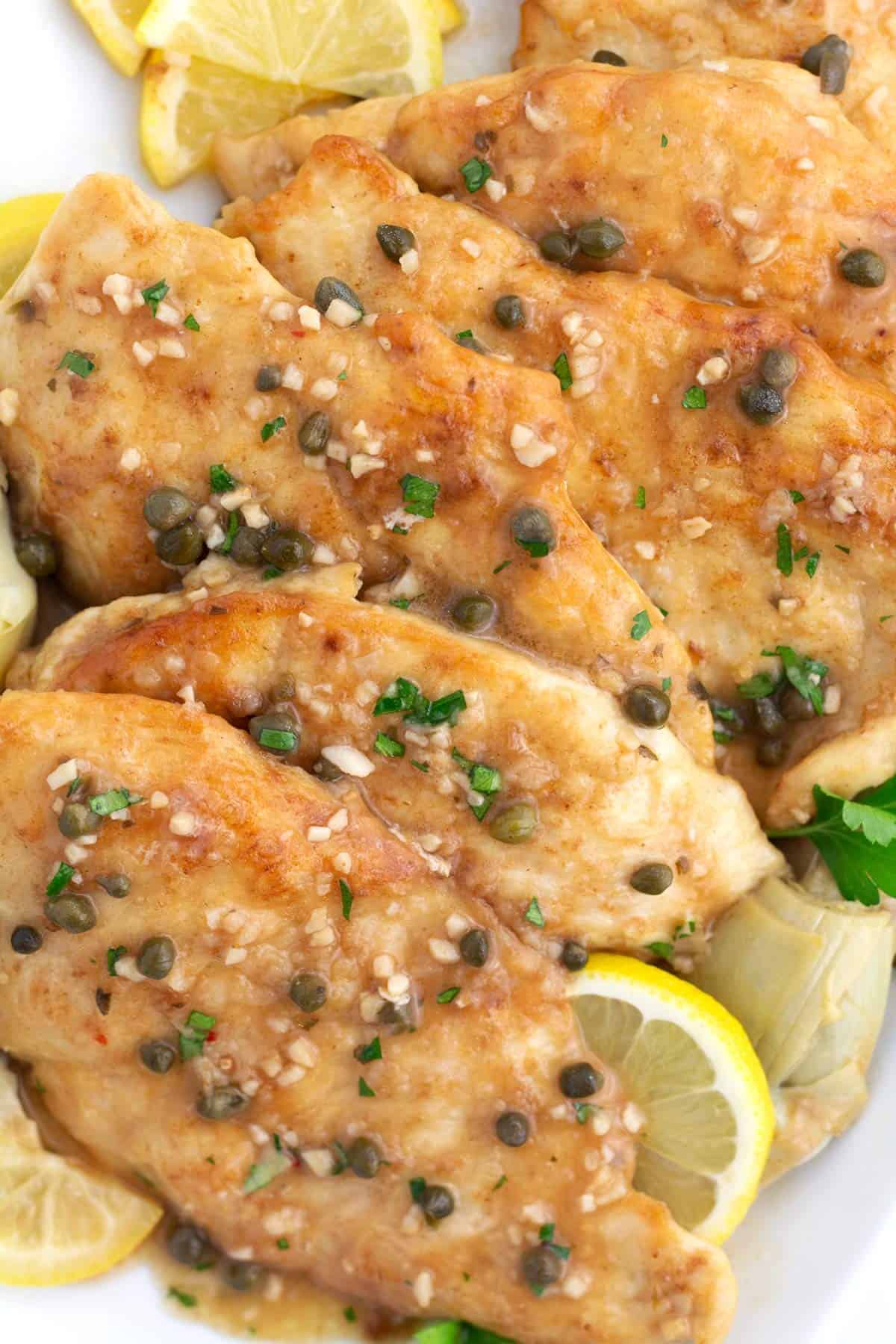 Overhead view of pieces of chicken piccata sprinkled with capers, parsley, garlic, and fresh lemon slices.