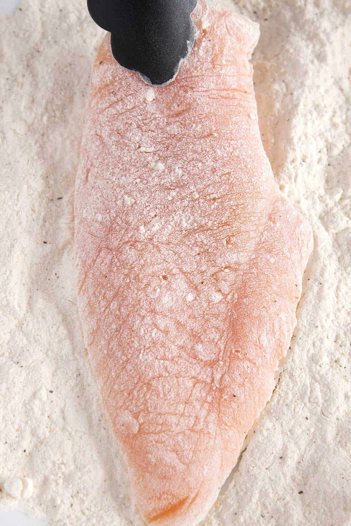 Raw chicken breast coated in a light layer of flour and sitting in a pile of more flour.