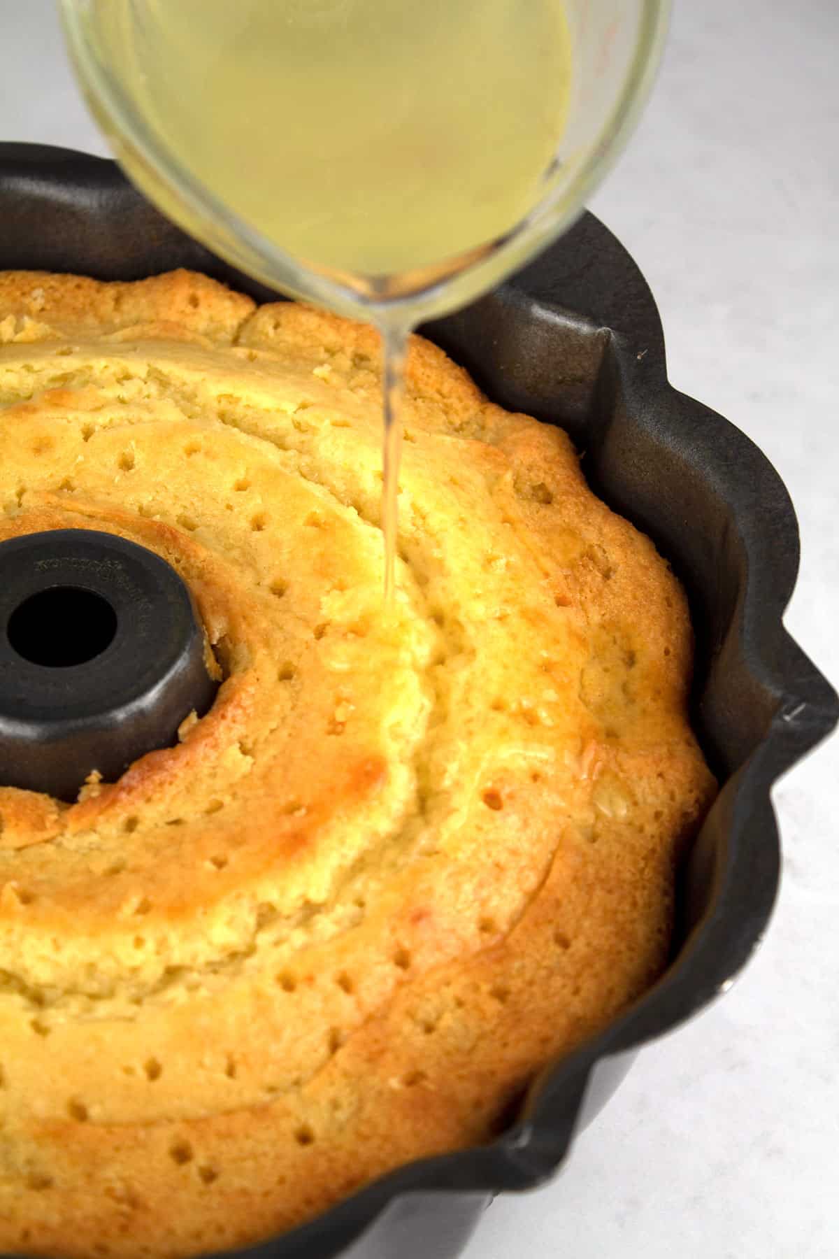 Pouring lemon simple syrup onto a bundt cake that has holes poked all over the top and is in a black bundt pan.