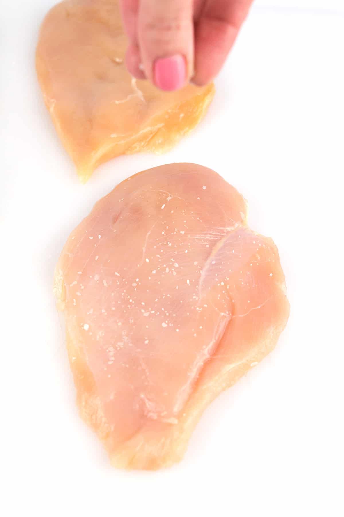 Hand sprinkling kosher salt onto a raw chicken breast on a white cutting board.