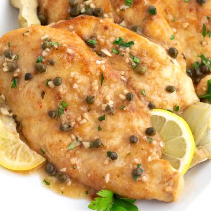 Pieces of chicken piccata on a white platter sprinkled with capers, parsley, garlic, and fresh lemon slices.