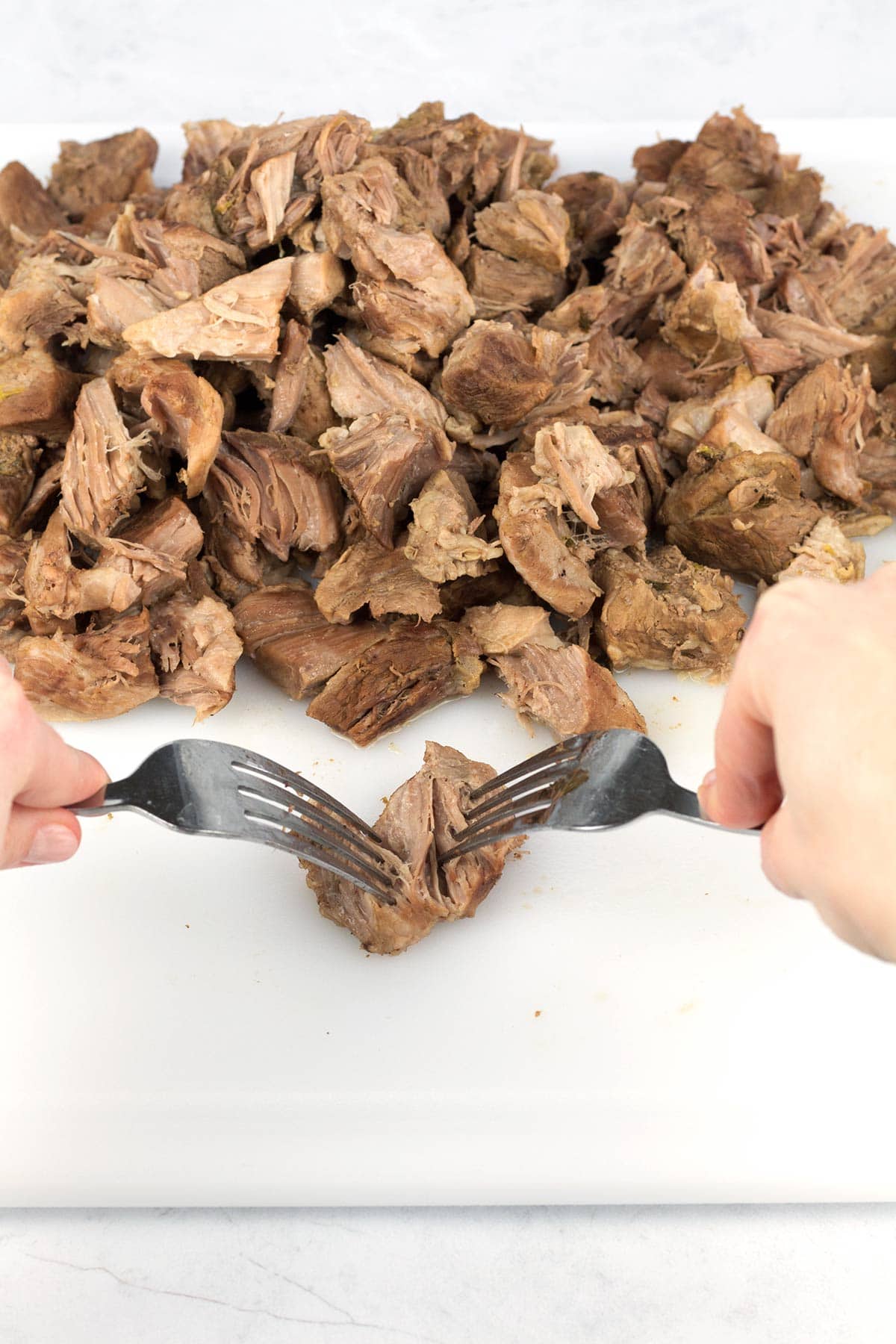 Shredding tender pork carnitas with two forks on a white cutting board.