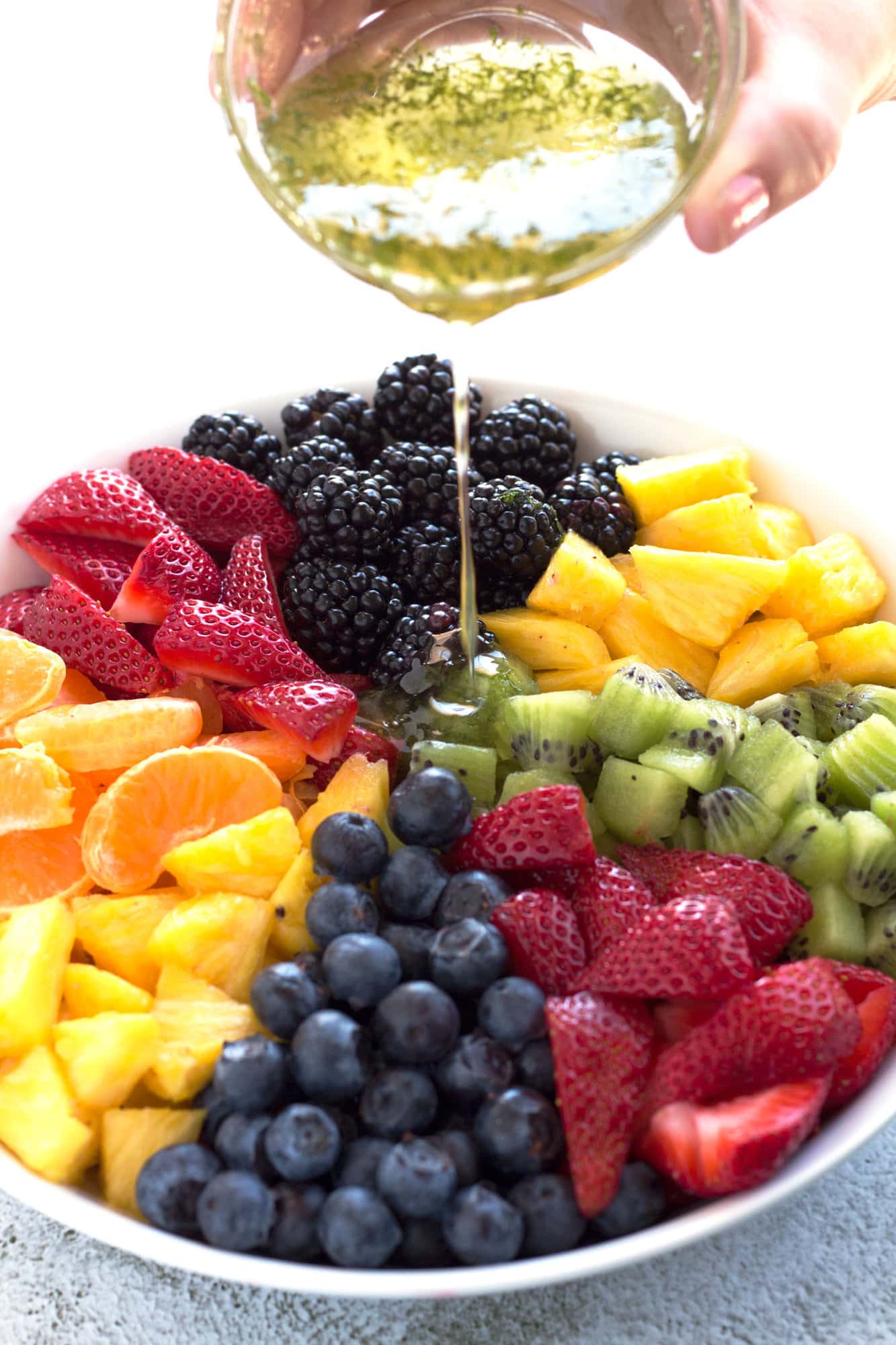 Hand pouring honey lime dressing on bowl of fruit salad.