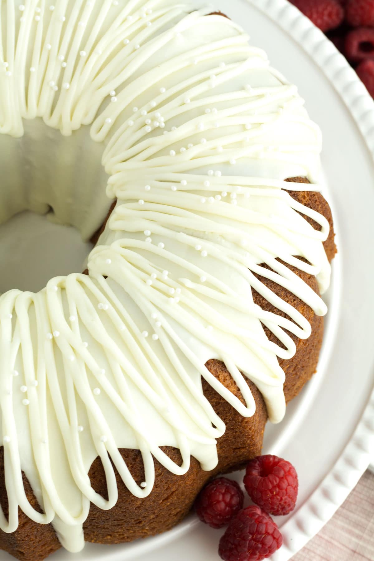 Whole decorated White Chocolate Raspberry Bundt Cake on a white cake plate with fresh raspberries.