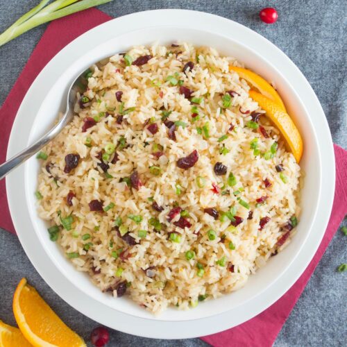 Orange rice with cranberries and scallions in white bowl on top of red and gray napkins.