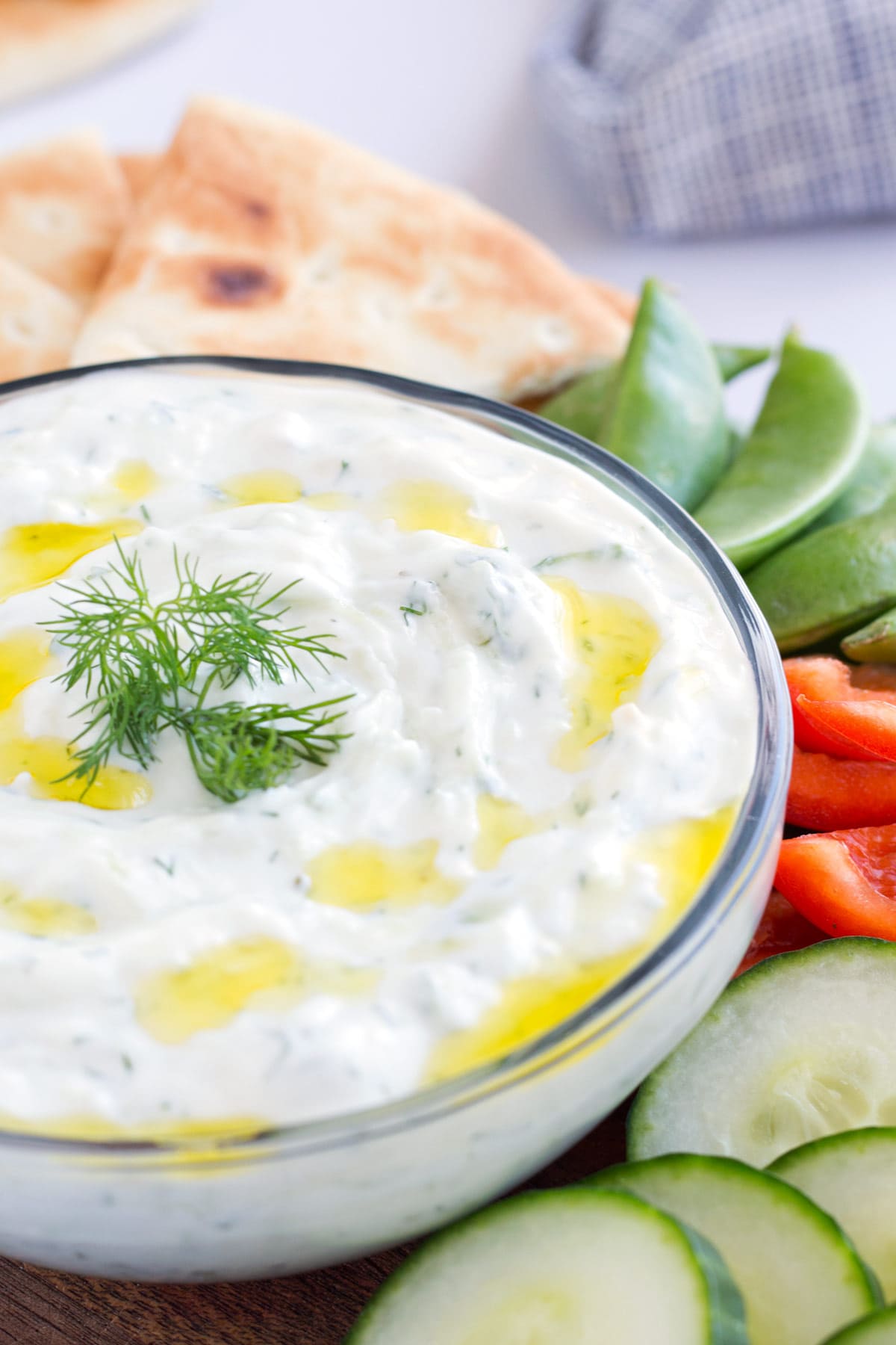 Glass bowl with creamy tzatziki sauce with olive oil on top and veggies on the side.