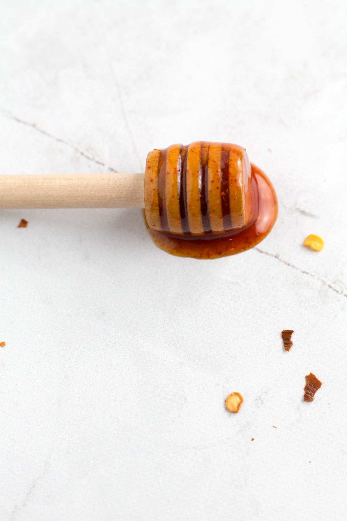 A honey dipper laying on the counter in a buddle hot honey sauce with chili pepper flakes sprinkled around.