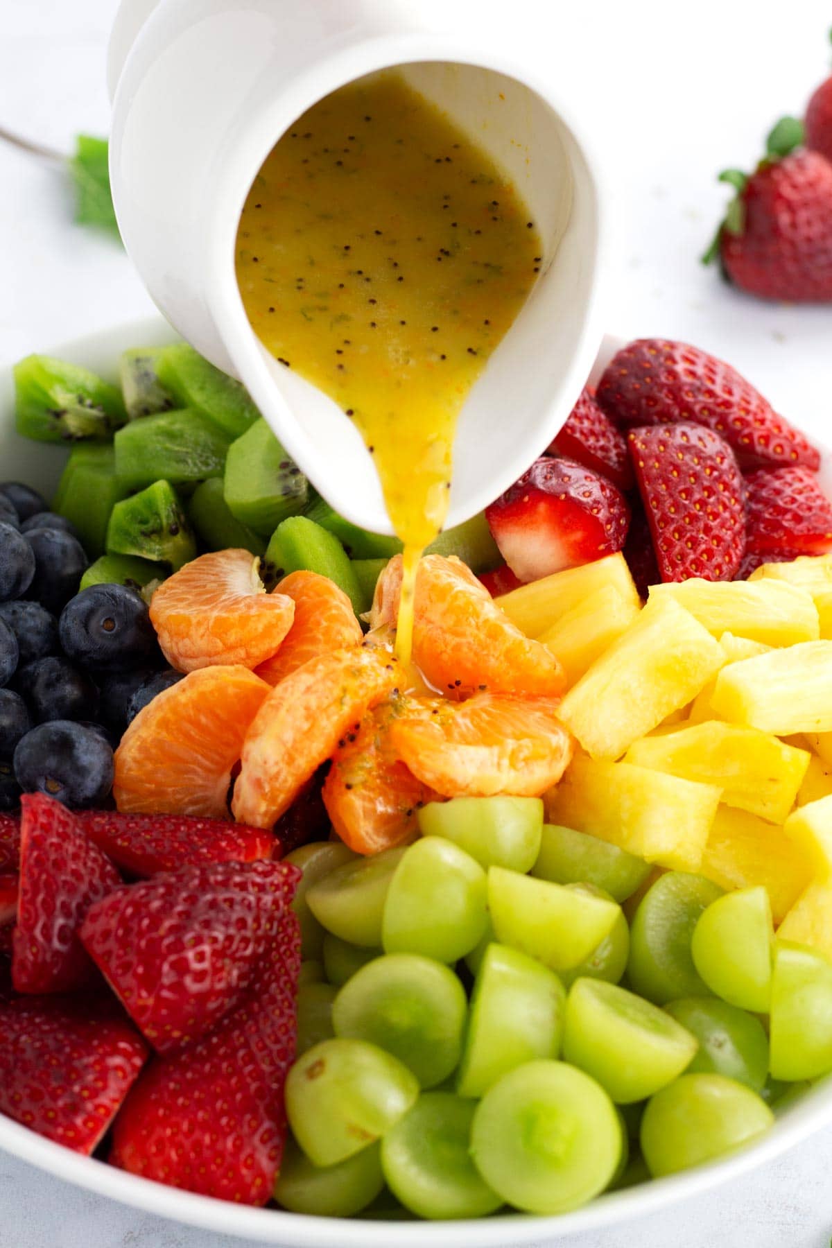 White jar pouring dressing for fruit salad onto a large bowl of fresh fruit.