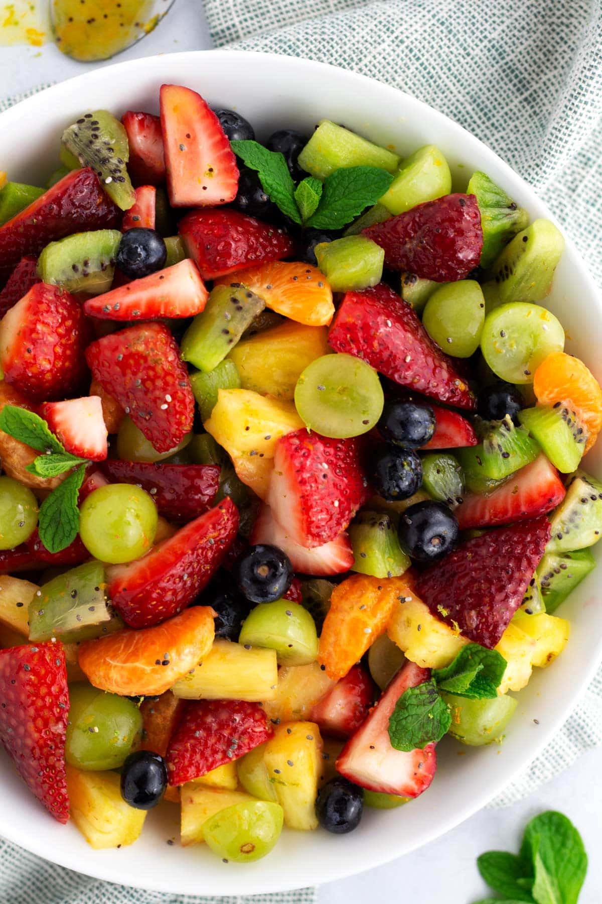Overhead view of a bowl of fruit salad covered in poppy seed dressing with a light green towel underneath.