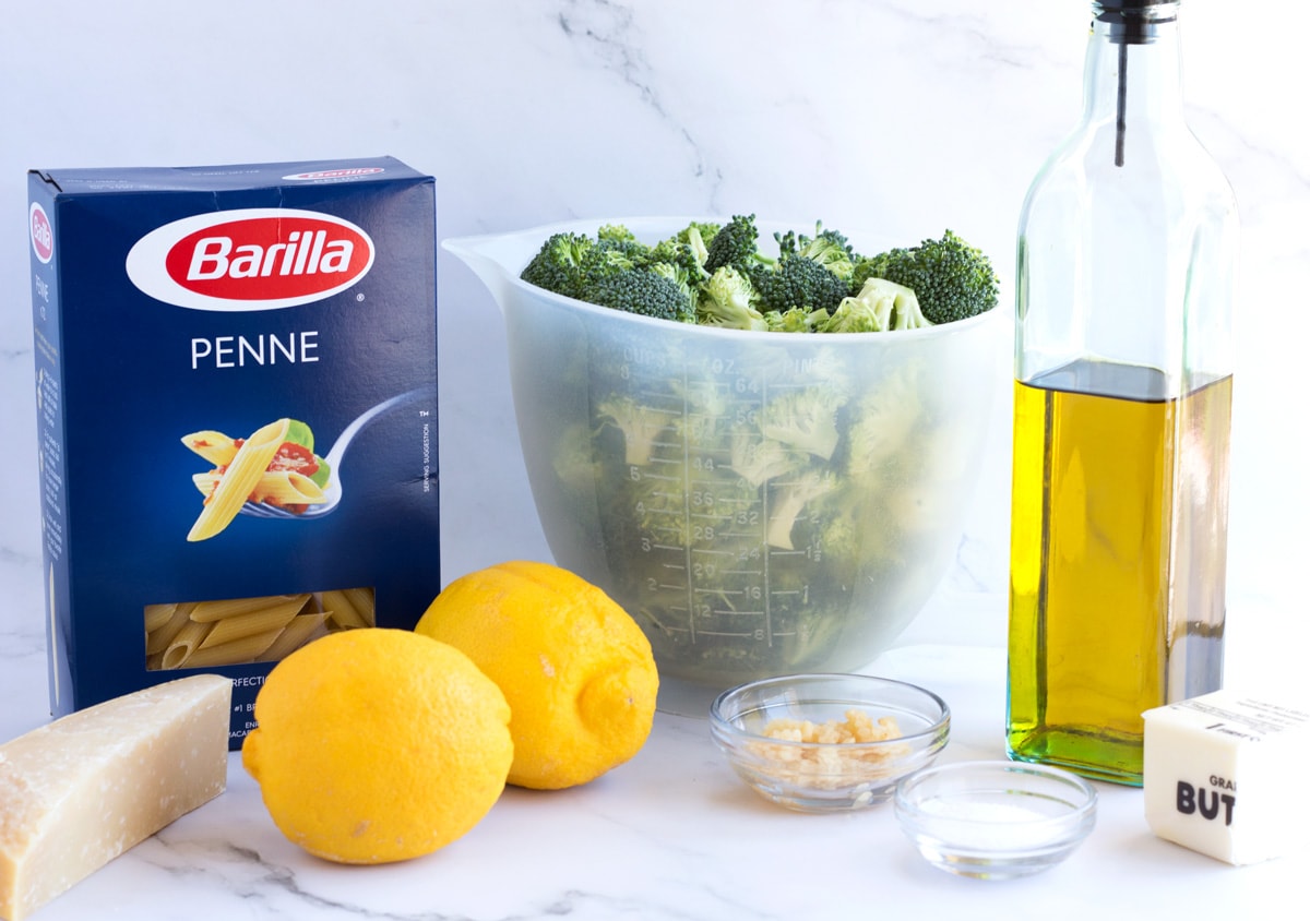 Pasta, broccoli, lemons, garlic, and parmesan cheese on a white marble countertop.