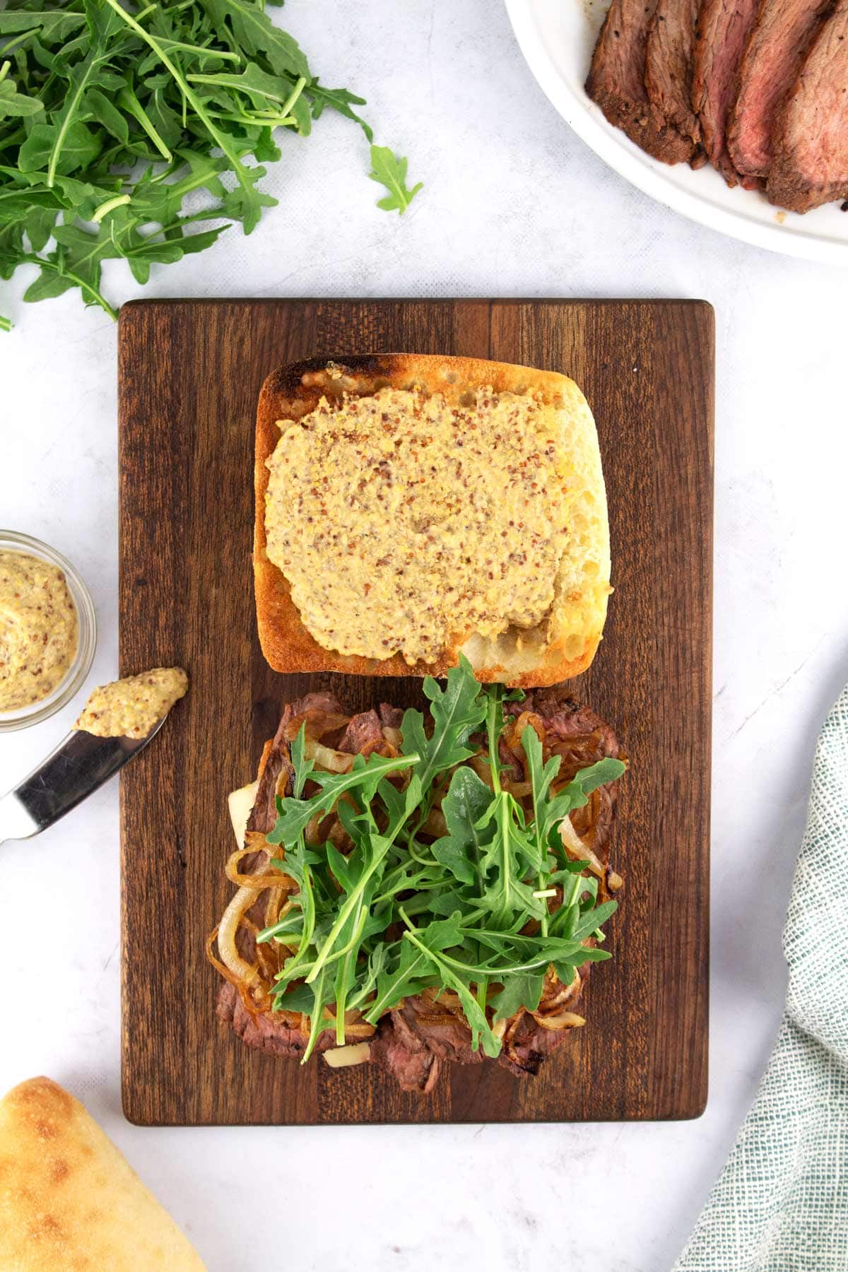 Overhead view of brown cutting board with open-face tri tip sandwich on top and extra ingredients on the sides.