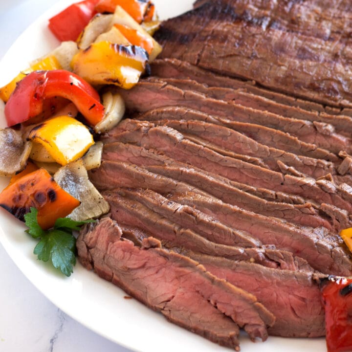 Medium rare sliced flank steak on white plate surrounded with grilled bell peppers and onions.