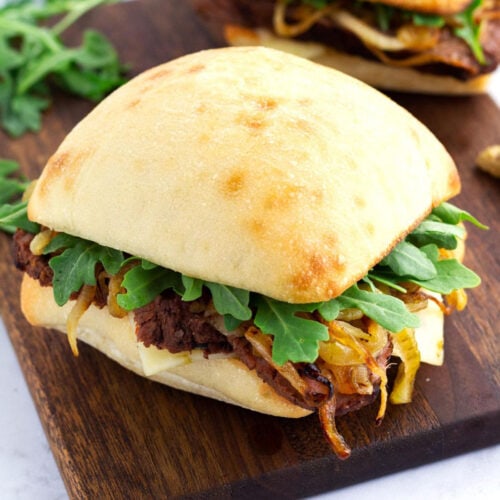 Tri tip steak sandwich on brown cutting board with onions, lettuce, and cheese sticking out of sandwich.