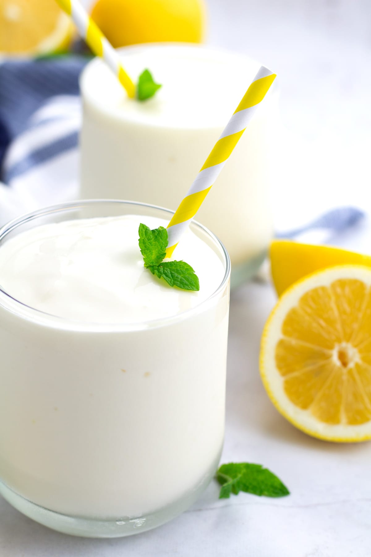 Two glasses of frozen lemonade with yellow straws, fresh mint, and fresh lemons around the glass.