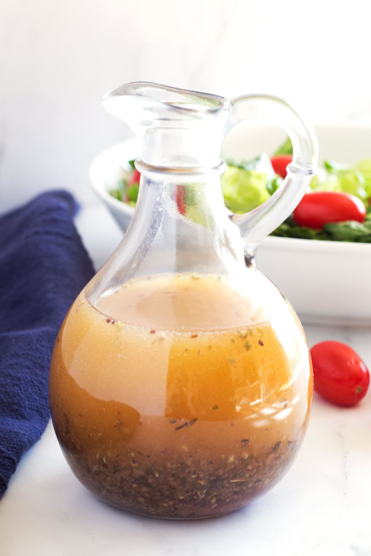 Straight view of a glass dressing bottle with Greek dressing inside and a bowl of salad in background.