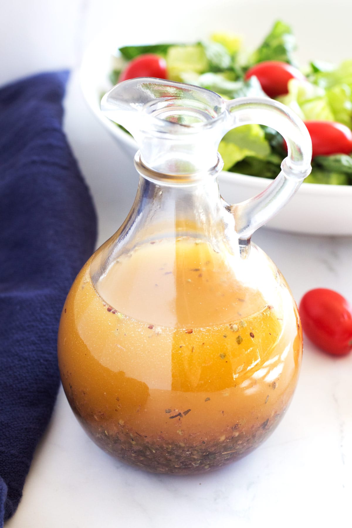 Clear jar of Greek vinaigrette with blue towel on side and green salad in background.