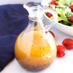 Clear jar of Greek vinaigrette dressing with blue towel on side and salad with tomatoes in background.