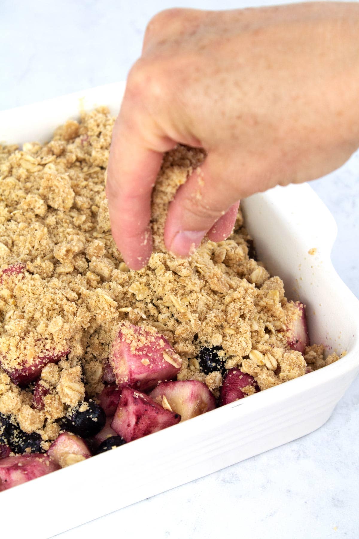 Hand sprinkling crumble on top of fruit in white baking pan.