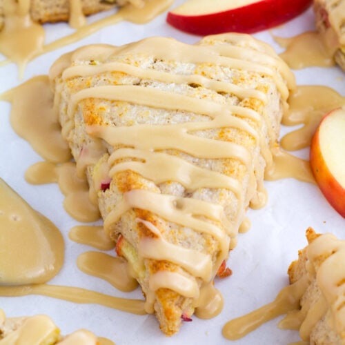 Apple scones on parchment paper with glaze drizzles and apple slices in background.