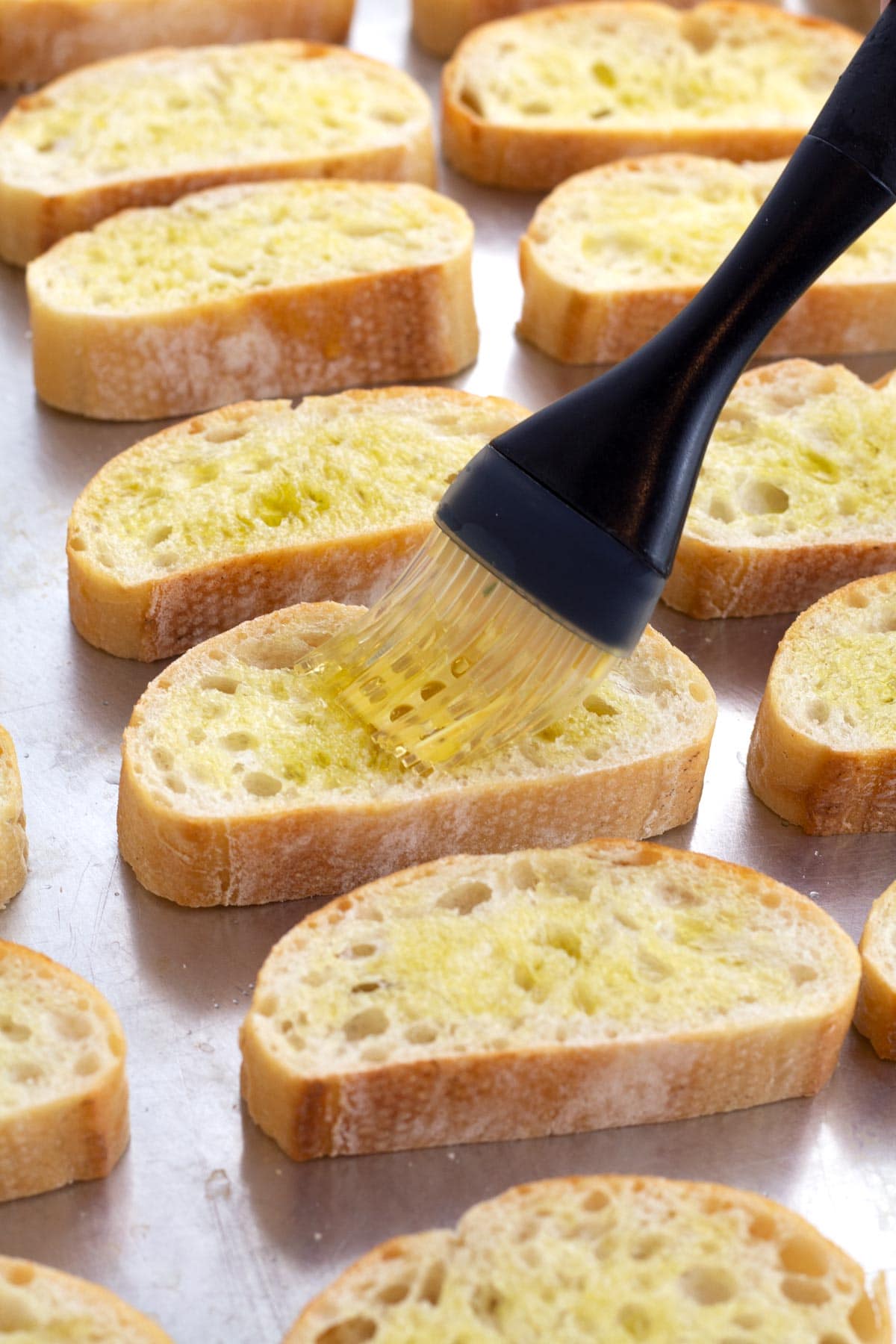 Silicone brush coating bread slices with olive oil on a silver sheet pan.