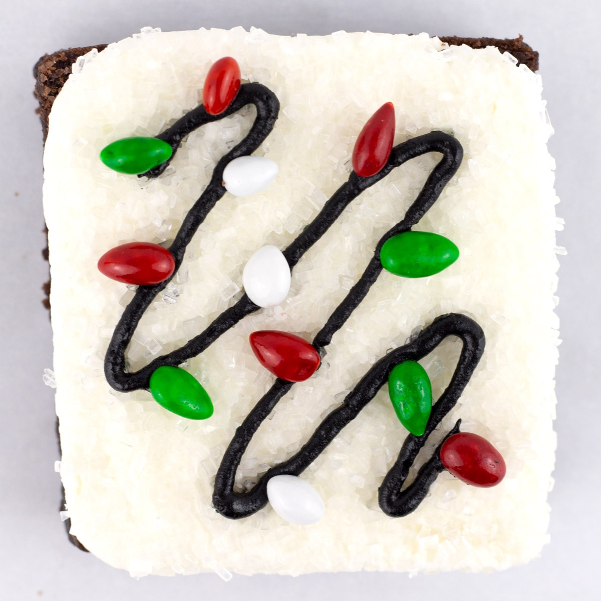 A square brownie decorated with Christmas lights using black frosting and chocolate covered sunflower seeds as the lights.