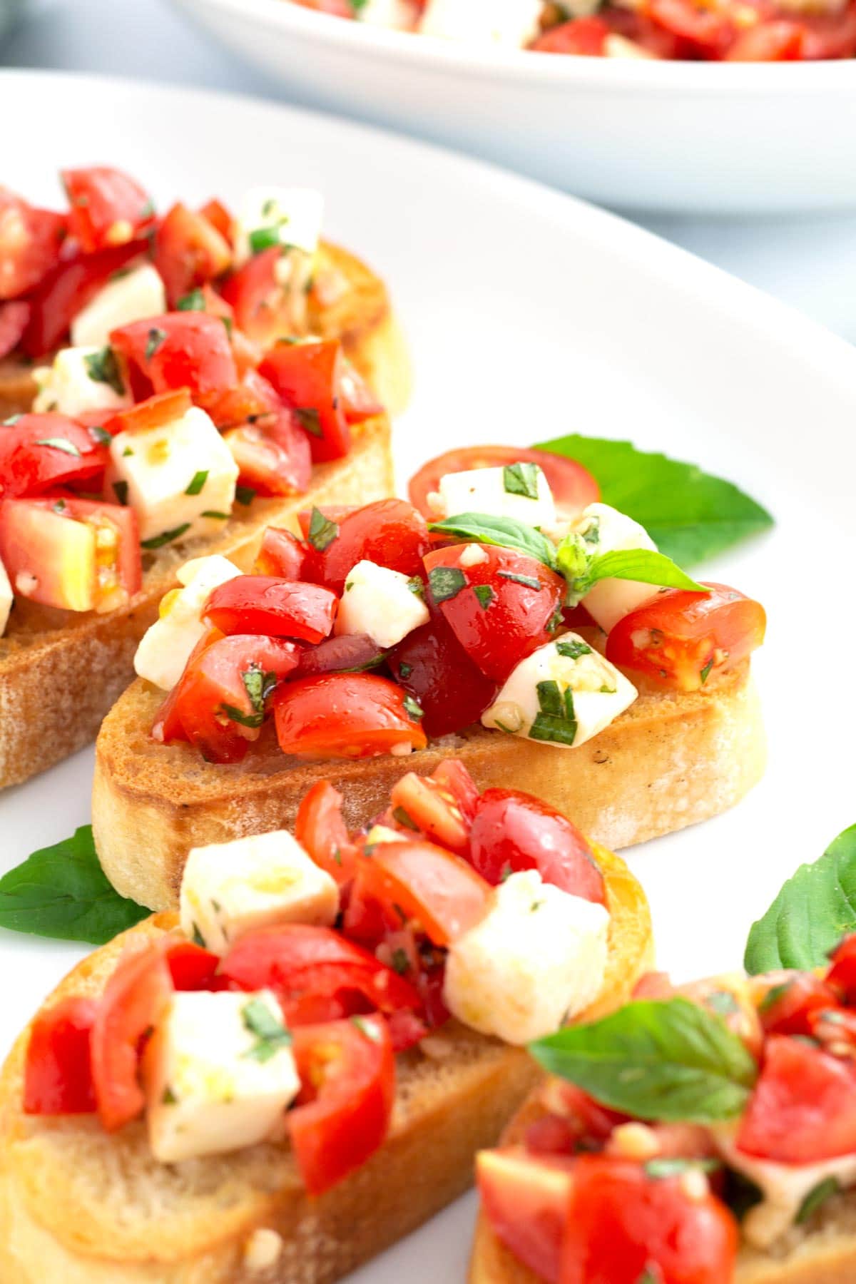 Five slices of bruschetta with mozzarella on a white tray with a serving bowl on the edge of the scene.
