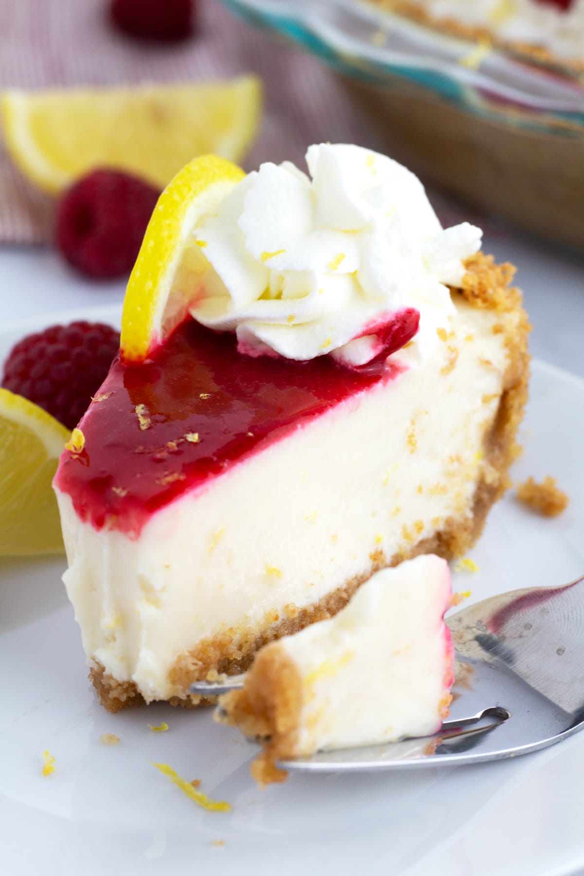 Slice of raspberry lemon pie on white plate with a fork on the side and lemon slices in the background.