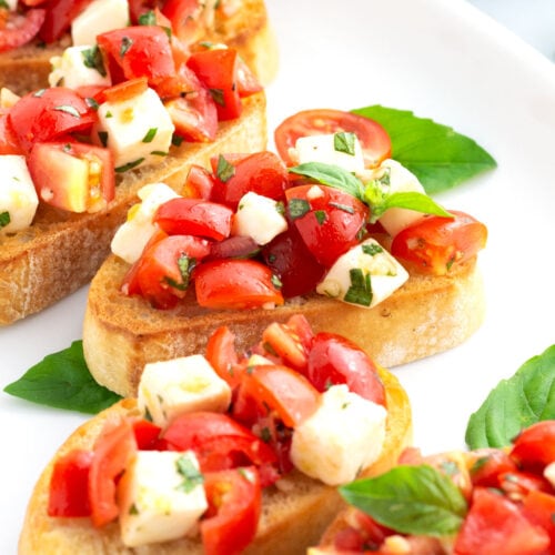 Mozzarella bruschetta served on a white tray with extra basil pieces in the scene.