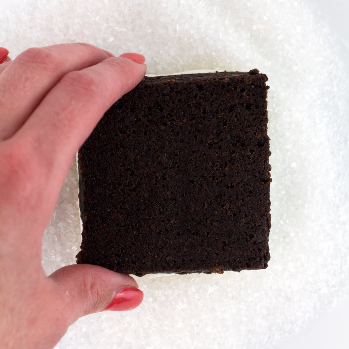 A hand dipping a frosted brownie into large sugar sprinkles.