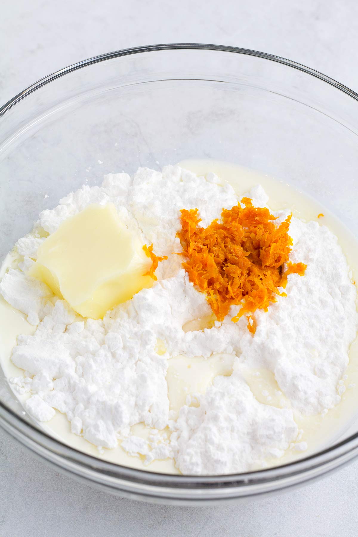 Glass bowl with powdered sugar, orange zest, and cream unmixed and a whisk resting in the bowl.