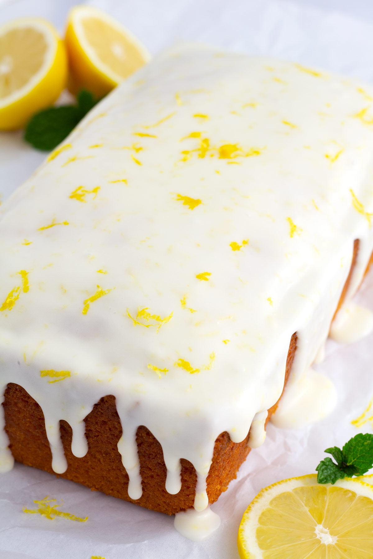 Whole lemon loaf on parchment paper dripping with lemon icing and sprinkled with fresh lemon zest.