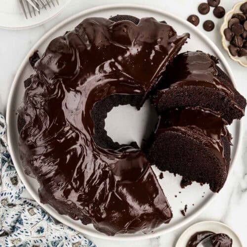 A chocolate bundt cake on a white platter with a slice cut out of it.