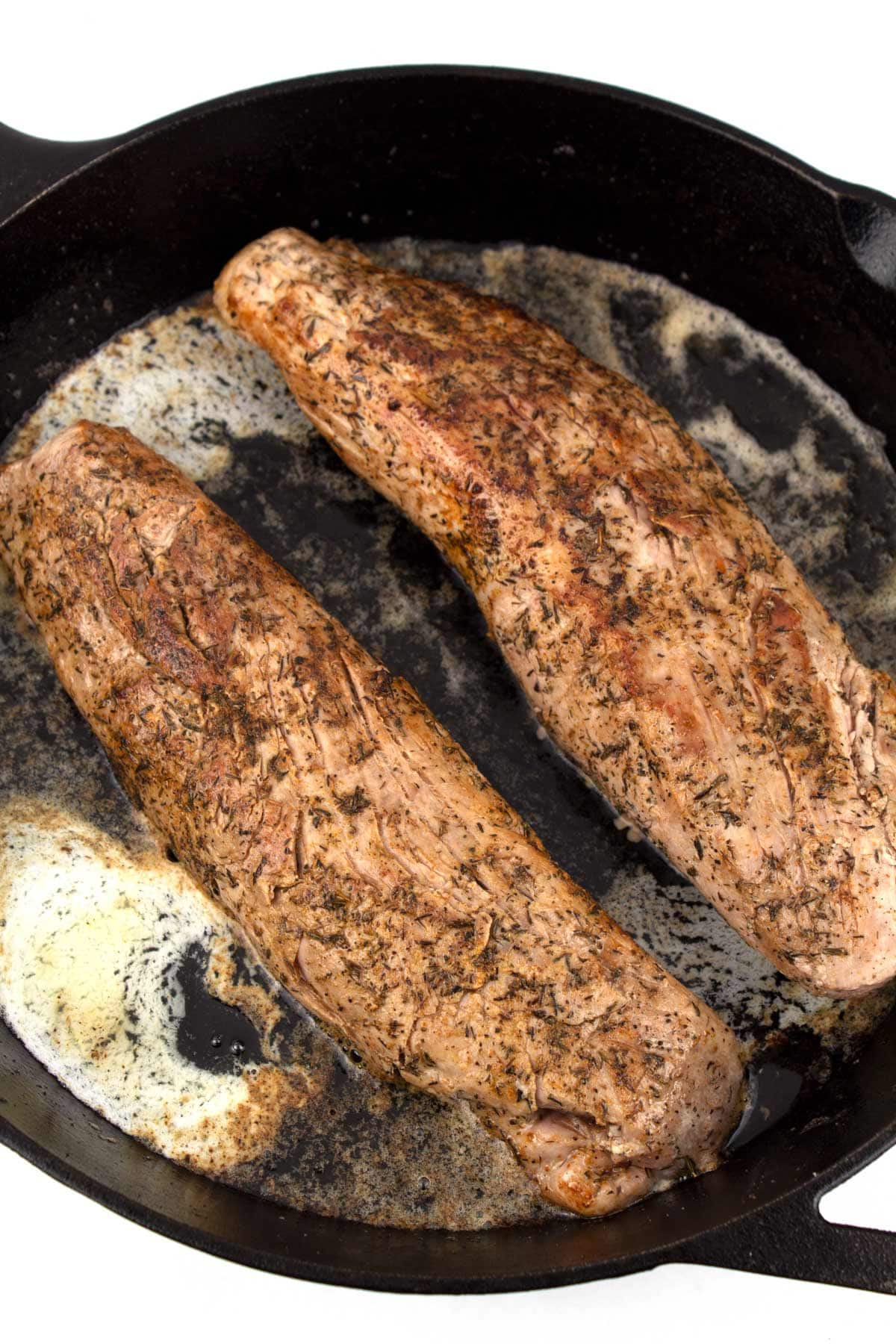 Two seared pork tenderloins in a cast iron skillet with honey and butter melting in the pan.