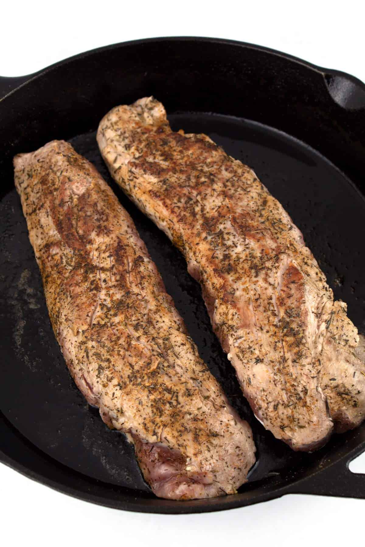 Two pork tenderloins searing in a cast iron skillet.