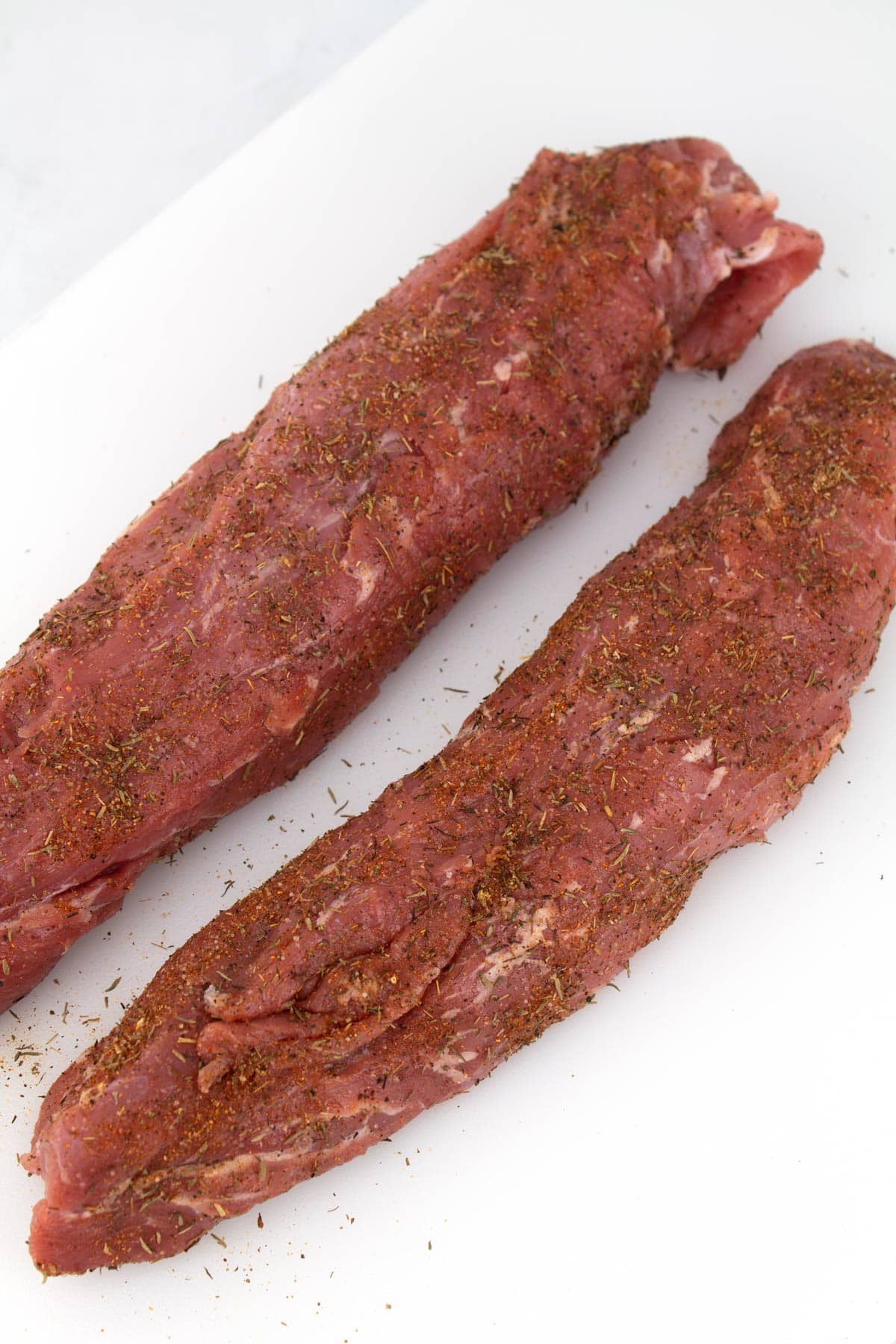 Two raw pork tenderloins sprinkled with seasoning on a white cutting board.