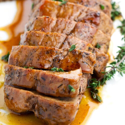 Sliced pork tenderloin on a white plate coated in sauce and sprinkled with fresh thyme.