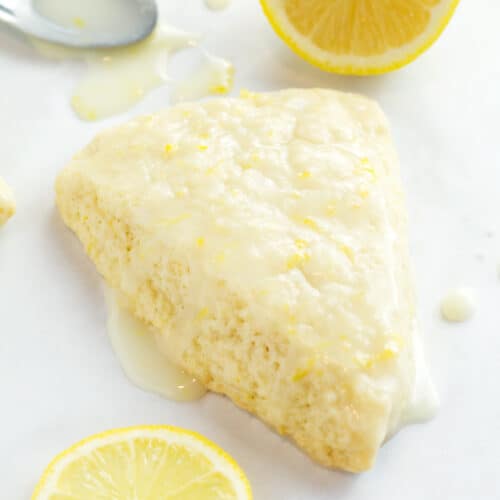 Lemon cream scone with lemon icing dripping around it and fresh lemon wedges in background.