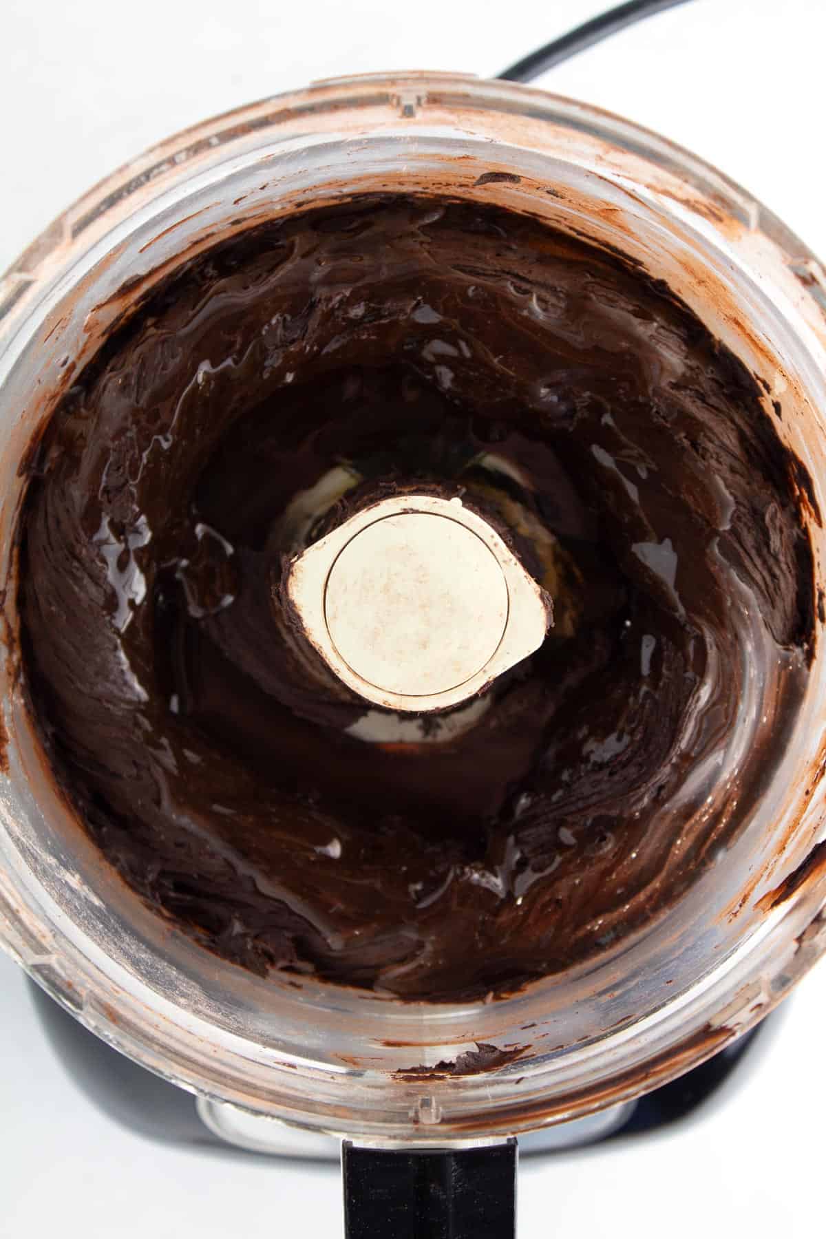 Chocolate frosting in a food processor with corn syrup poured on top.