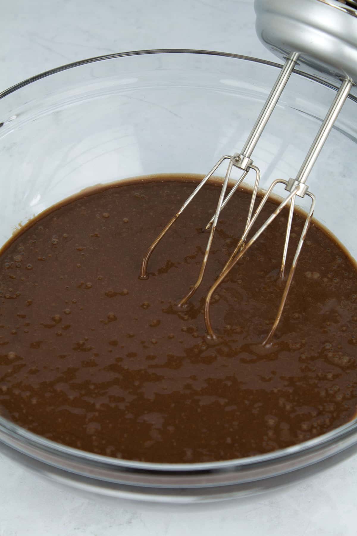 Finished chocolate cake batter in a clear glass bowl with hand mixers resting in the back.