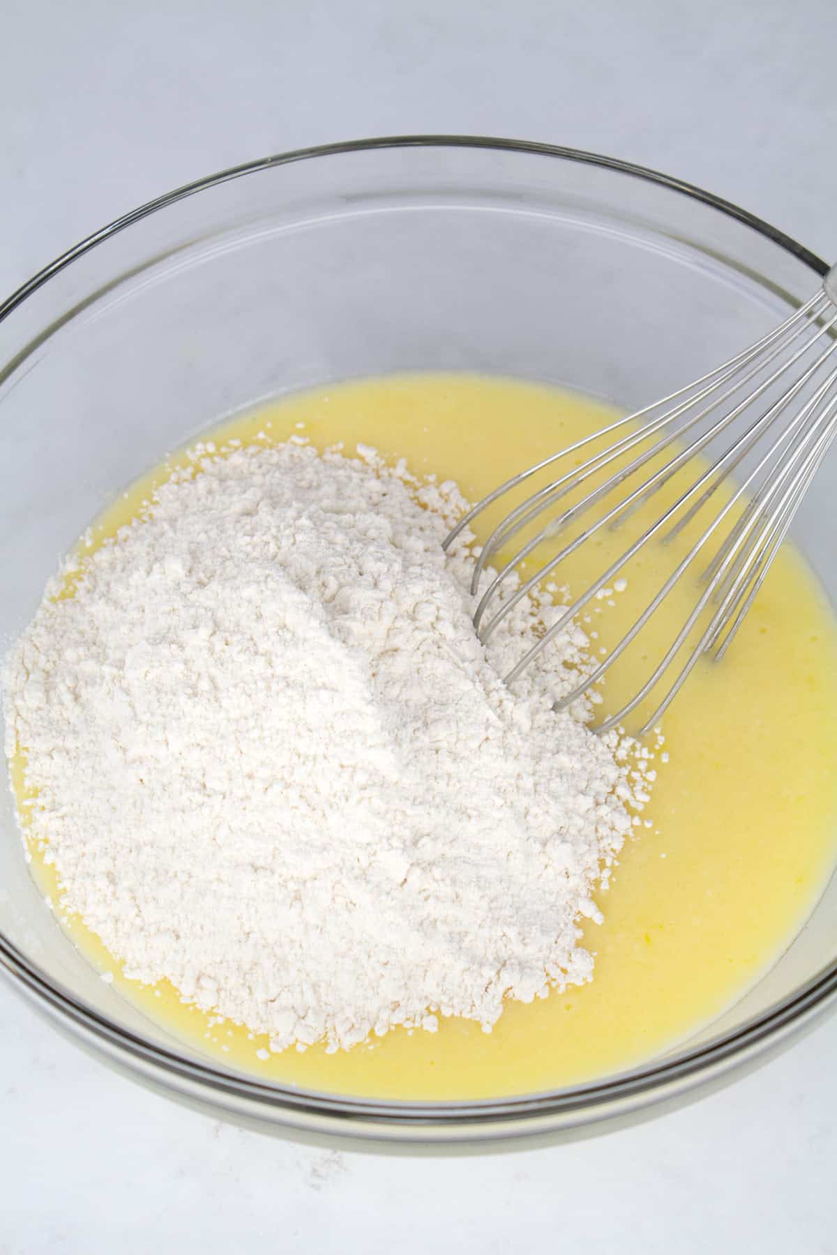 Flour mixture added on top of wet ingredients in a glass bowl with a whisk sitting in the back of the bowl.