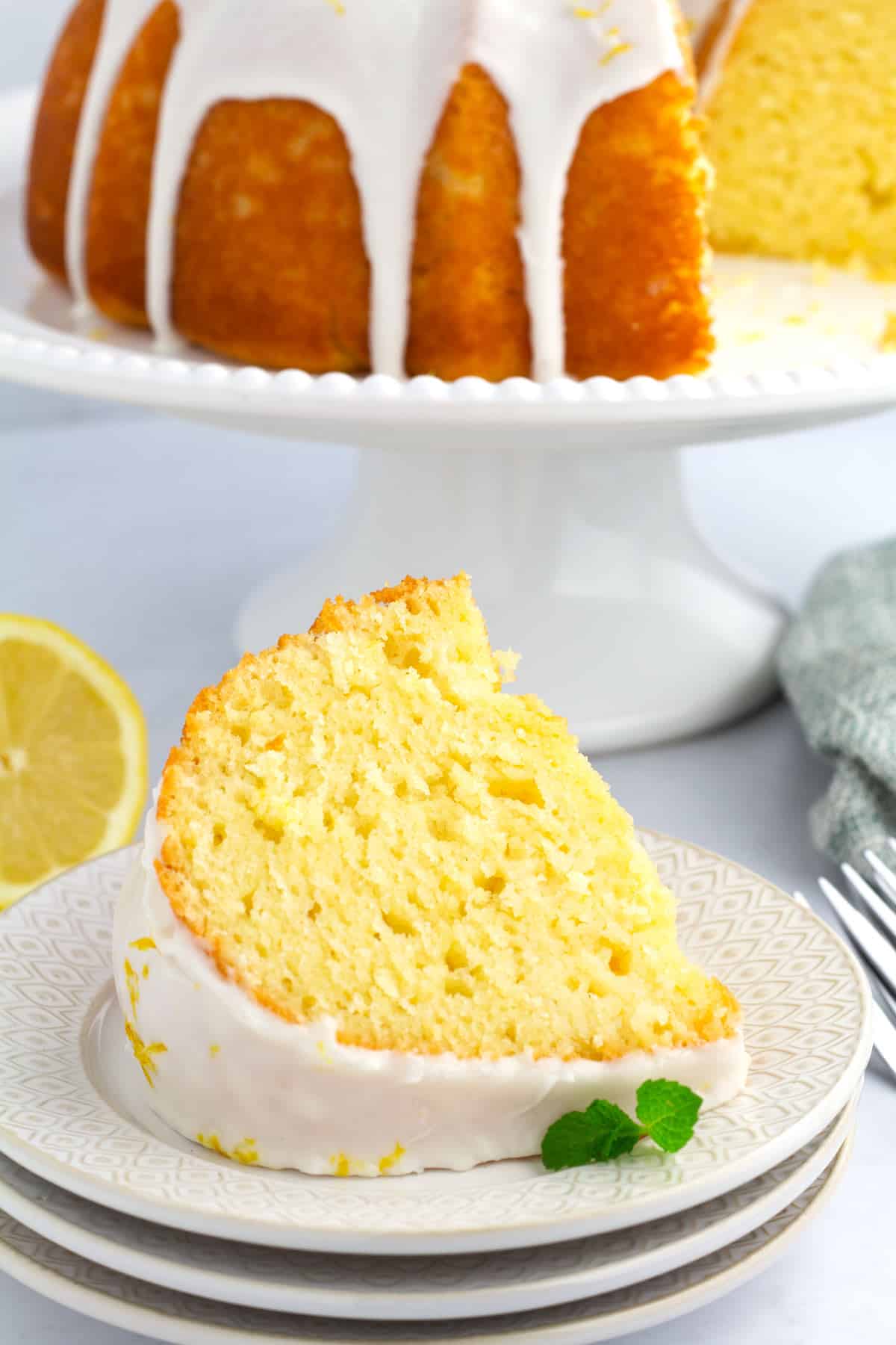 Slice of lemon cake on a plate with the full bundt cake and lemon slices in the background.