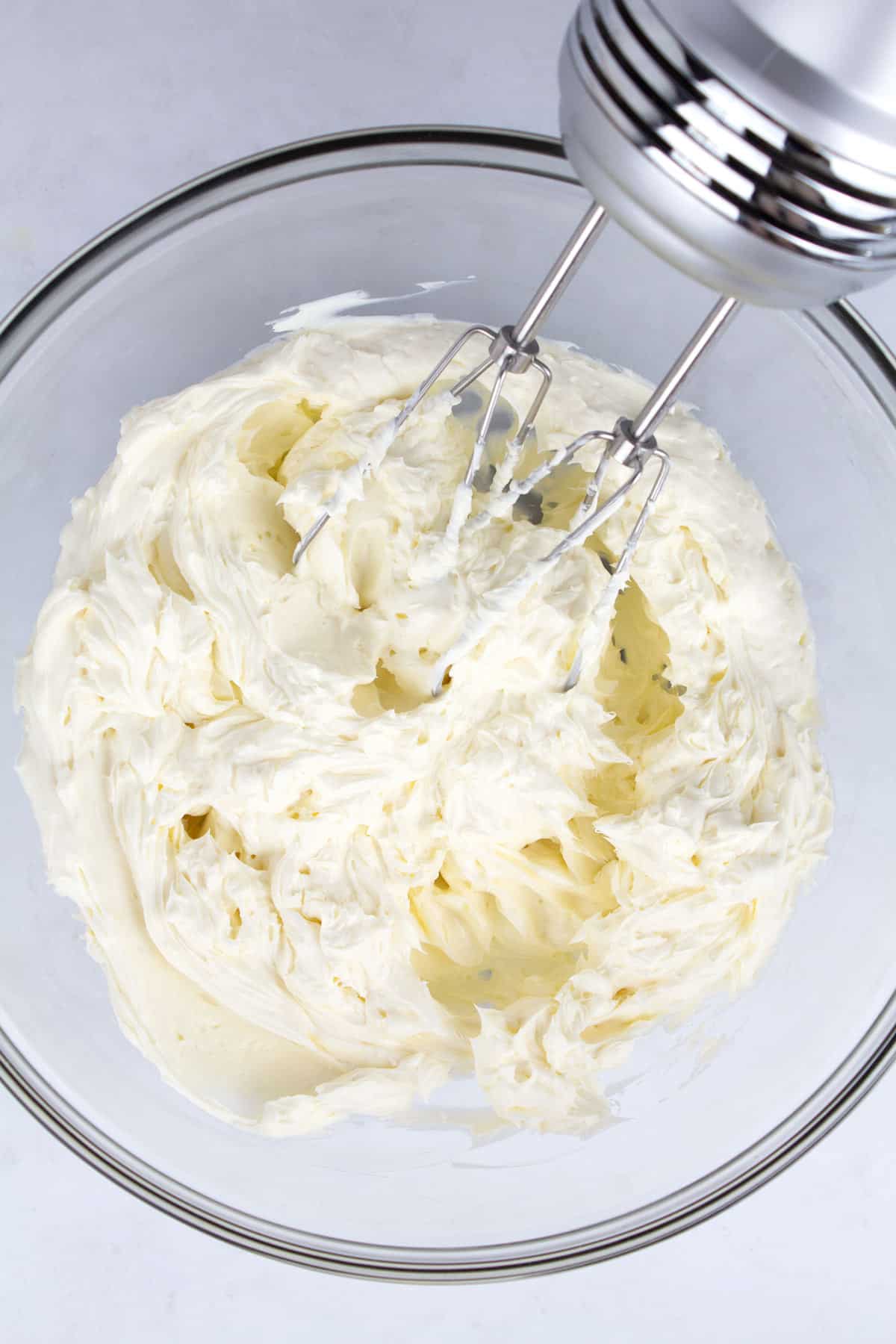 An overhead view of beaten cream cheese in a clear glass bowl with a hand mixer.