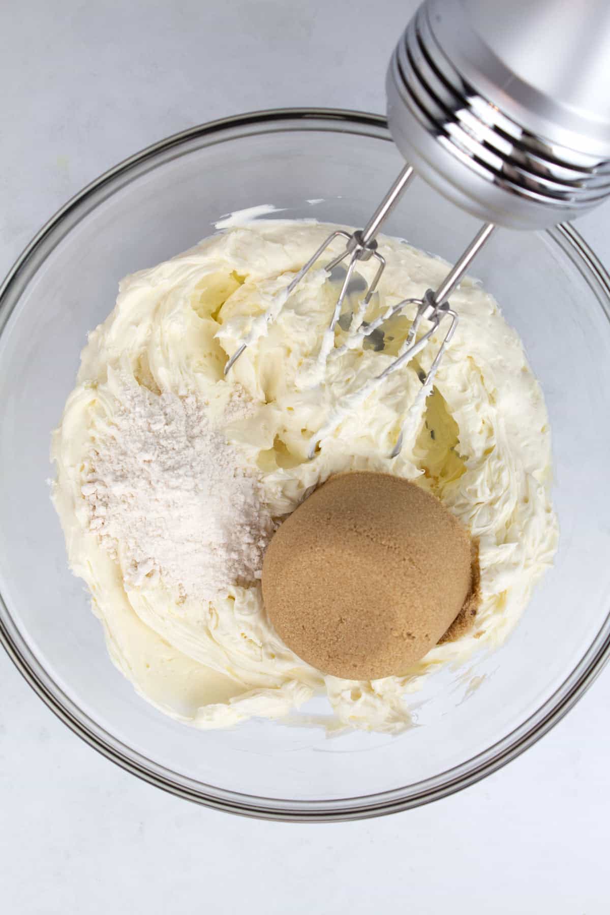 An overhead view of cream cheese, sugar, and flour in a clear glass bowl with a hand mixer.