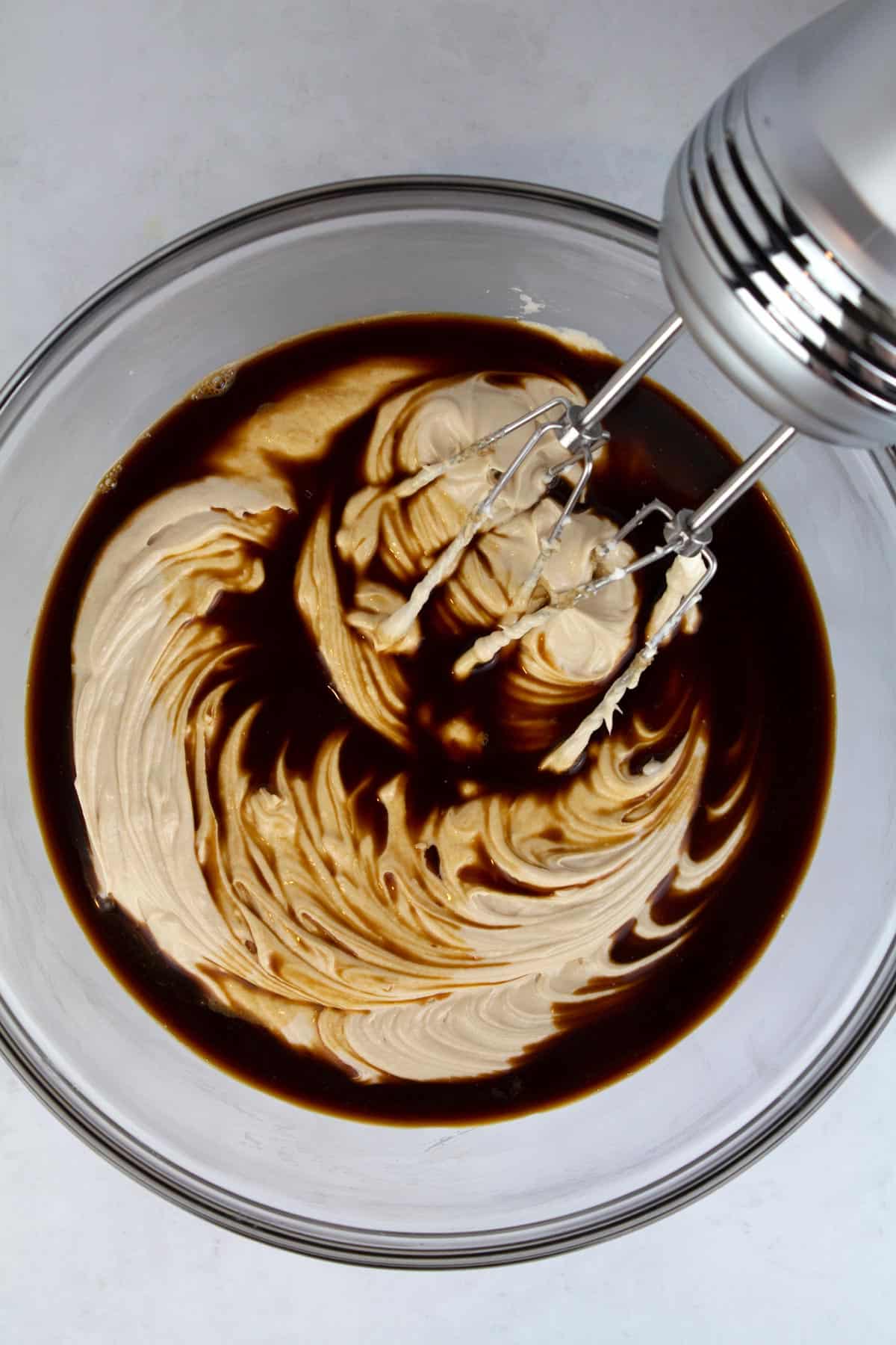An overhead view of cheesecake batter in a clear glass bowl with espresso added on top and a hand mixer sitting in the back.