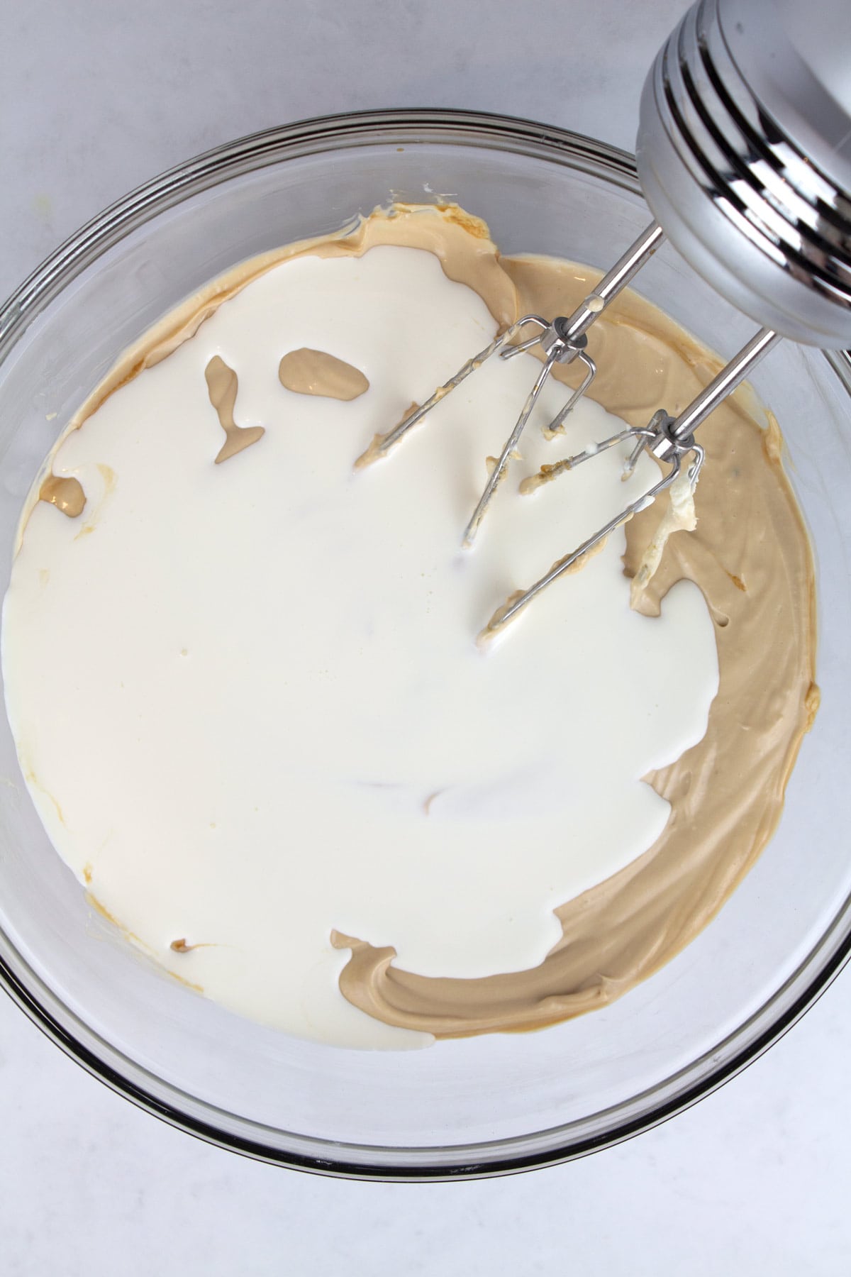 An overhead view of cheesecake batter in a clear glass bowl with cream added on top and a hand mixer in the back.