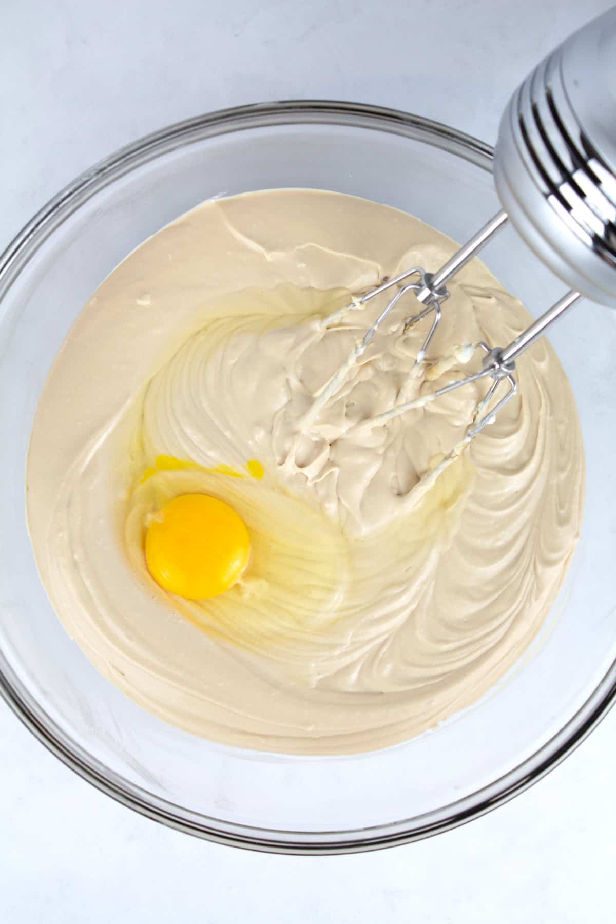 An overhead view of cheesecake batter with a raw egg on top in a clear glass bowl with a hand mixer resting in the bowl.